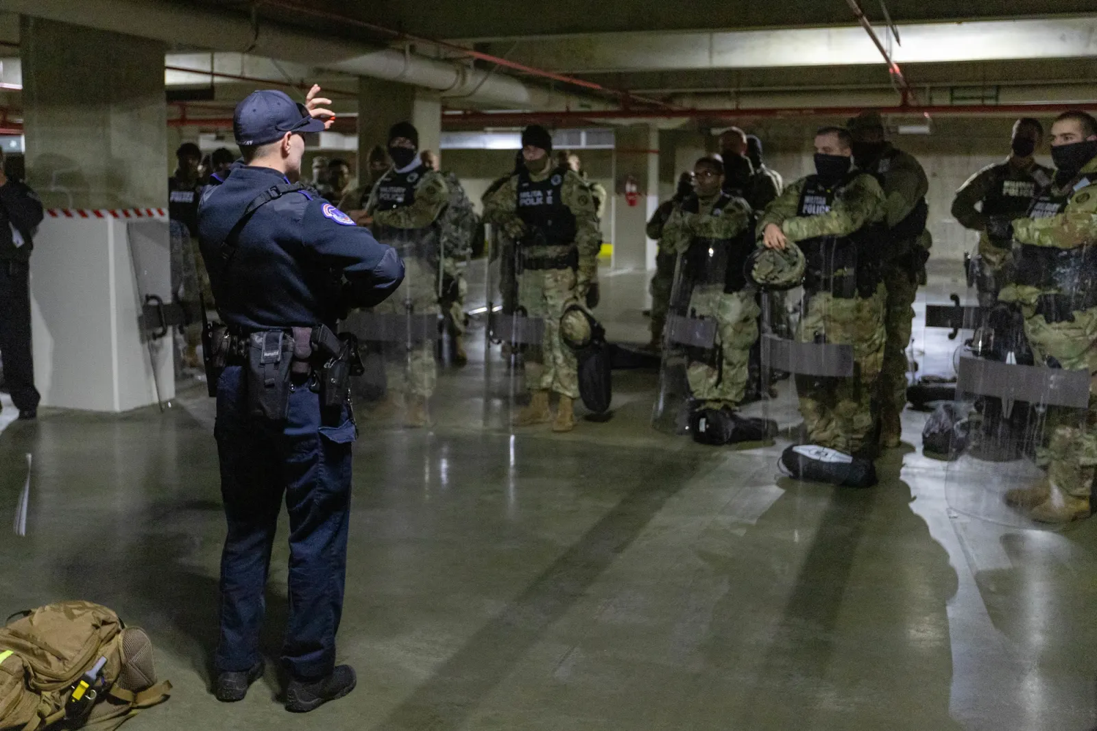 Capitol Police officer seen from behind, in a dark blue uniform and a loaded duty belt, facing National Guard military police with shields